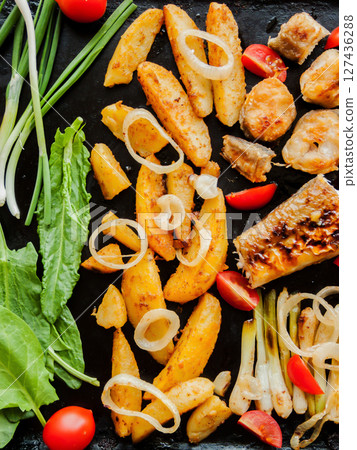 Oven pan with sliced baked potatoes, fish steaks and vegetables with onion on the table. Homemade healthy dinner with potatoes and seafood. 127436288