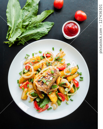 Plate with baked potatoes, piece of fish, vegetables and herbs on the table with tomato sauce. Dinner served with fish and chips meal. Plate with baked potatoes, piece of fish, vegetables and herbs on the table with tomato sauce. Dinner served with fish and chips meal. 127436301