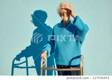Portrait, shadow and disability with a senior woman on a blue wall background while holding a mobility walker outdoor. Health, handicap and fashion with a mature female standing outside alone 127436453