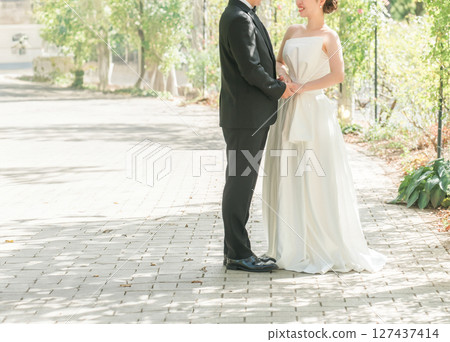 A couple in dresses walking hand in hand along a tree-lined path in a park (wedding, wedding photo, bride and groom) A couple in dresses walking hand in hand along a tree-lined path in a park (wedding, wedding photo, bride and groom) 127437414