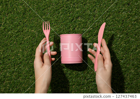 Pink tin can and plastic cutleries in hands on green background, top view Pink tin can and plastic cutleries in hands on green background, top view 127437463