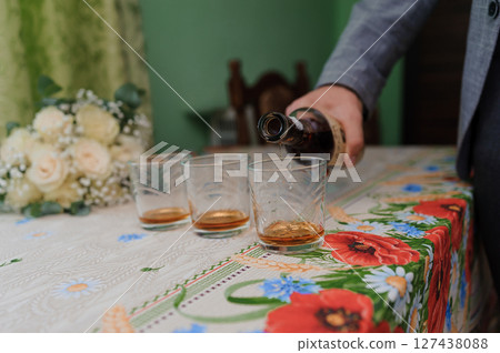 A man pours amber liquor from a dark glass bottle into three clear glasses on a table covered with a floral tablecloth during a wedding celebration 127438088