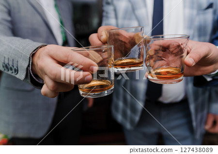 Three well-dressed men clinking rocks glasses filled with amber liquid, likely whiskey, celebrating a joyful occasion with camaraderie. Three well-dressed men clinking rocks glasses filled with amber liquid, likely whiskey, celebrating a joyful occasion with camaraderie. 127438089