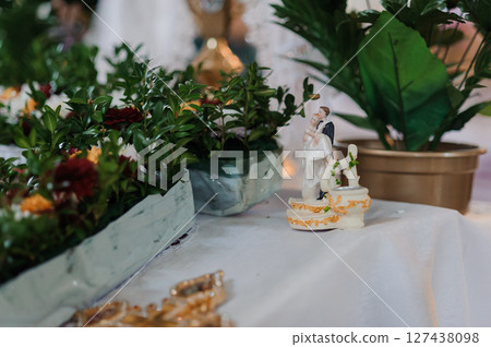 Wedding cake topper figurine of a couple with a dog, set among lush greenery and floral arrangements on a white tablecloth Wedding cake topper figurine of a couple with a dog, set among lush greenery and floral arrangements on a white tablecloth 127438098