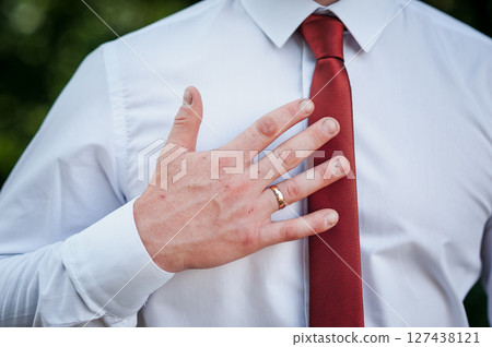 Man in white shirt and red tie placing hand over heart, golden wedding ring visible, solemn expression, outdoor setting Man in white shirt and red tie placing hand over heart, golden wedding ring visible, solemn expression, outdoor setting 127438121