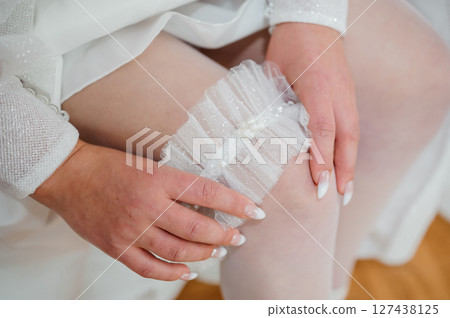 A close-up shot of a bride delicately holding a shimmering white lace garter in her hands, preparing for a wedding ceremony detail, with soft skin tones and delicate fabrics. A close-up shot of a bride delicately holding a shimmering white lace garter in her hands, preparing for a wedding ceremony detail, with soft skin tones and delicate fabrics. 127438125