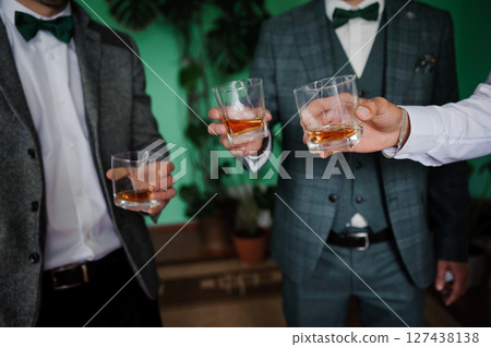 Three groomsmen in stylish tweed suits clinking glasses with whiskey in a modern interior with green plants and a blurred background. Three groomsmen in stylish tweed suits clinking glasses with whiskey in a modern interior with green plants and a blurred background. 127438138