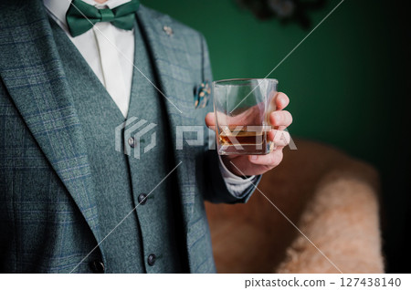 Stylish man in a green tweed suit with a bowtie holds a glass of whiskey, enjoying a drink in an elegant setting. 127438140