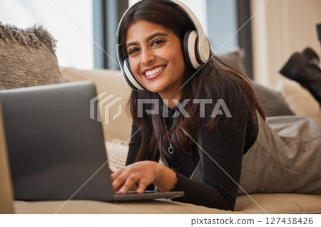 Headphone, woman and relax on laptop, happy and listening to music, podcast or watch a movie on the sofa in home. Portrait, young girl and happy with streaming subscription app, audio or social media 127438426
