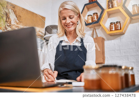 Laptop, small business and senior woman writing notes while doing inventory or online marketing. Happy, smile and elderly entrepreneur in a honey store working on admin or shop sales with a computer. Laptop, small business and senior woman writing notes while doing inventory or online marketing. Happy, smile and elderly entrepreneur in a honey store working on admin or shop sales with a computer. 127438478