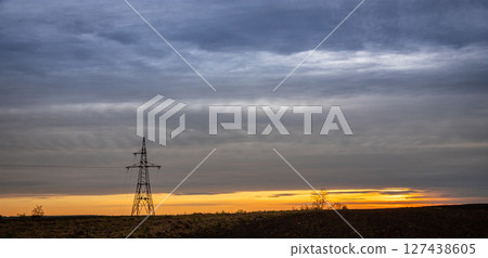 Sunset over a vast landscape with a lone power tower silhouetted against the colorful evening sky near a rural area Sunset over a vast landscape with a lone power tower silhouetted against the colorful evening sky near a rural area 127438605