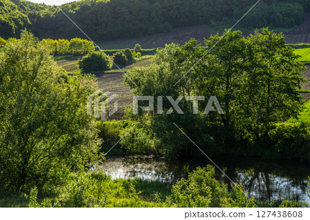 Serene countryside landscape with lush trees calm water and rolling fields during a sunny afternoon Serene countryside landscape with lush trees calm water and rolling fields during a sunny afternoon 127438608
