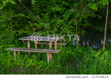 Wooden picnic table and benches set in a serene green setting beside a calm stream during late afternoon Wooden picnic table and benches set in a serene green setting beside a calm stream during late afternoon 127438614
