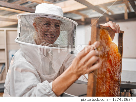 Beekeeper, honey and manufacturing with frame, honeycomb and suit for safety in workshop or farm. Portrait of agriculture employee or animal farmer happy, working and farming natural resource Beekeeper, honey and manufacturing with frame, honeycomb and suit for safety in workshop or farm. Portrait of agriculture employee or animal farmer happy, working and farming natural resource 127438684