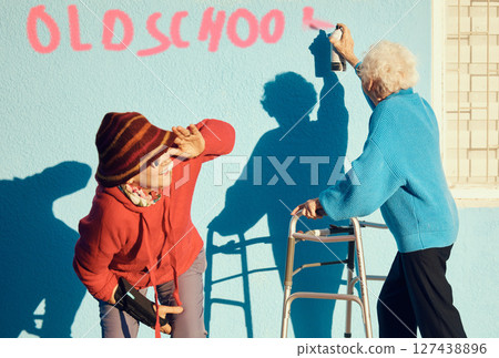 Senior women, friends and spray paint for humor, graffiti and street art on wall of nursing home building. Old people breaking the law with illegal activity outdoor 127438896