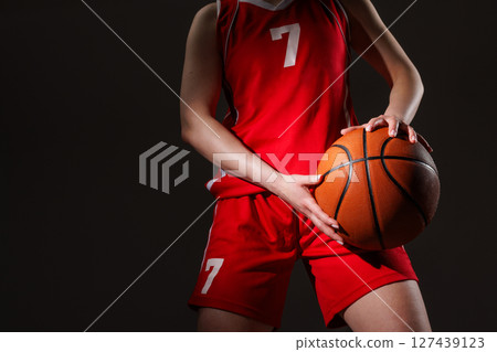 Confident female basketball player in red uniform holding ball in front of her, ready to compete under dramatic studio lighting. 127439123
