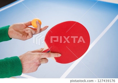 Close-up of hands holding red table tennis paddle and orange ball, ready to serve on a blue ping pong table. Close-up of hands holding red table tennis paddle and orange ball, ready to serve on a blue ping pong table. 127439176