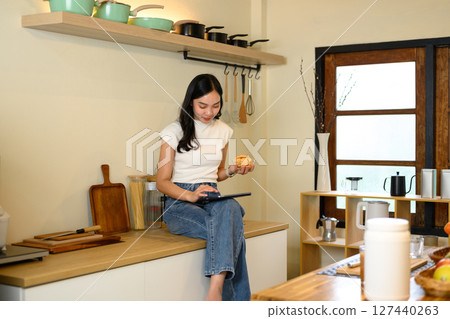 Young woman using digital tablet and enjoying a snack while sitting on kitchen counter Young woman using digital tablet and enjoying a snack while sitting on kitchen counter 127440263