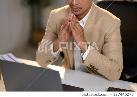 Close up of male executive in beige suit deep in thought while working on a laptop at desk 127440272