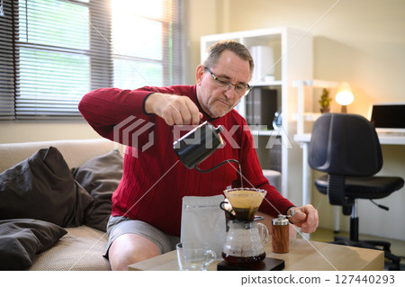 Senior man in red sweater making pour over coffee at home with a focused expression Senior man in red sweater making pour over coffee at home with a focused expression 127440293