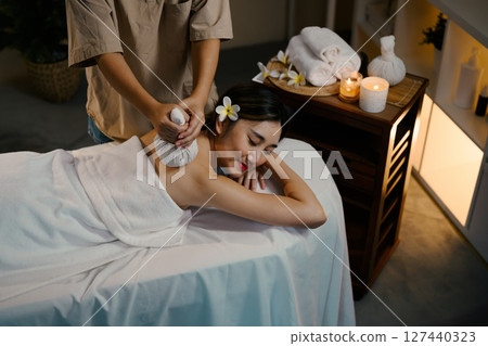 Young woman receiving traditional herbal compress massage at spa with relaxing candlelit ambiance Young woman receiving traditional herbal compress massage at spa with relaxing candlelit ambiance 127440323