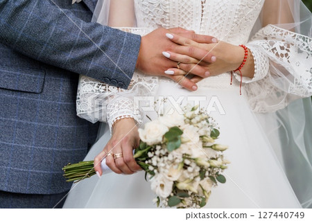 Romantic Wedding Close-up of Hands with Red String and Bouquet, Stylish Groom, Lace White Dress, Soft Focus Floral Backdrop. 127440749