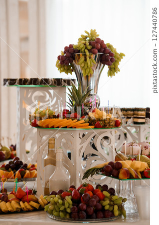 Elegant Fruit Buffet Display: Gourmet Assortment of Grapes, Pineapple, Strawberries, Melons, and Fresh Fruits in Crystal Vases, White Decor Elegant Fruit Buffet Display: Gourmet Assortment of Grapes, Pineapple, Strawberries, Melons, and Fresh Fruits in Crystal Vases, White Decor 127440786