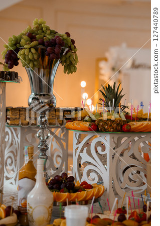 Luxurious Fruit Table Setting with Grapes, Pineapple, Oranges, Silver Vases, and Elaborate White Decorations for an Elegant Event. Luxurious Fruit Table Setting with Grapes, Pineapple, Oranges, Silver Vases, and Elaborate White Decorations for an Elegant Event. 127440789