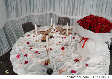 Romantic Dinner Setup with Red Roses, Candles and Wine: Cozy Intimate Evening Scene, Soft Lighting and White Petal Decorations 127440801