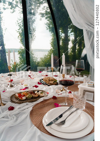 Romantic Dinner Setup in Glass Dome Tent Overlooking Lake. Candles, Red Wine, Rose Petals, Table Setting, Scenic View, White Linens, Cozy Atmosphere Romantic Dinner Setup in Glass Dome Tent Overlooking Lake. Candles, Red Wine, Rose Petals, Table Setting, Scenic View, White Linens, Cozy Atmosphere 127440802