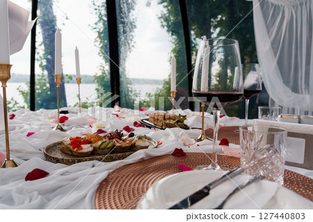 Romantic Lakeside Dinner Setup with Wine, Rose Petals, and Appetizers in a Glass-Walled Structure, Soft Lighting, White Tablecloth Romantic Lakeside Dinner Setup with Wine, Rose Petals, and Appetizers in a Glass-Walled Structure, Soft Lighting, White Tablecloth 127440803