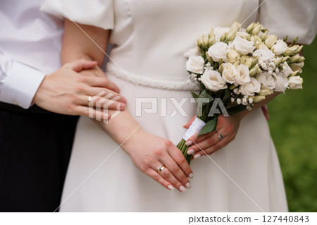 Romantic Wedding Day Close-Up: Bride and Groom Hands with Rings, White Bouquet, Elegant Dress, Outdoor Greenery, Symbolic Love and Commitment. 127440843