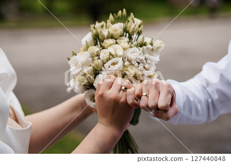 Wedding Rings and Bouquet Close-Up, Hands Holding Flowers, Green Background, Romantic Gold Bands, Symbolic Love, Outdoor Ceremony Shot, Soft Focus. 127440848