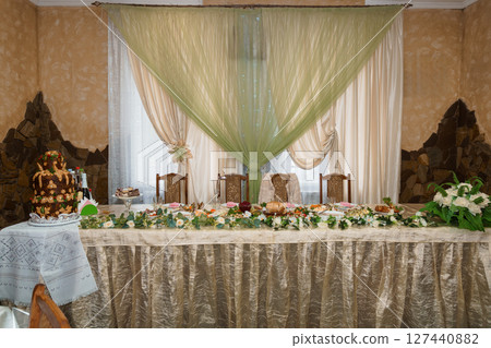 Luxurious Wedding Table Setting with Floral Decorations and Cake in Elegant Room Interior Featuring Beige Walls, Green Drapes, and Stone Accents. Luxurious Wedding Table Setting with Floral Decorations and Cake in Elegant Room Interior Featuring Beige Walls, Green Drapes, and Stone Accents. 127440882