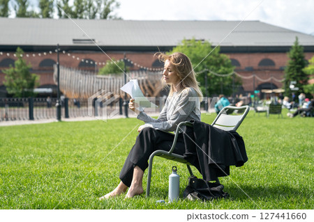 Uncomfortable woman fanning with sheets of paper like fan while sitting on chair outside on lawn. 127441660