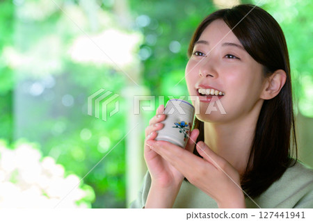 一位年輕女子在室內享受下午茶時光，周圍環繞著自然的夏日陽光，面帶微笑，手裡拿著茶杯 127441941