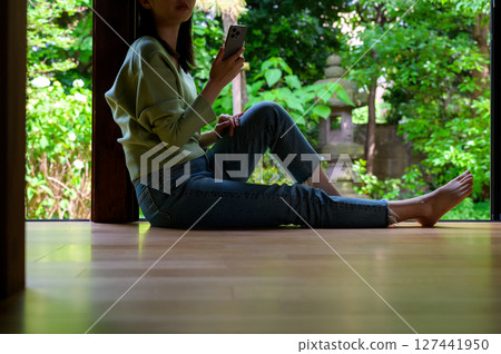 A woman stretches her legs out on the veranda where natural light pours in and operates her smartphone during a calm summer evening. A woman stretches her legs out on the veranda where natural light pours in and operates her smartphone during a calm summer evening. 127441950