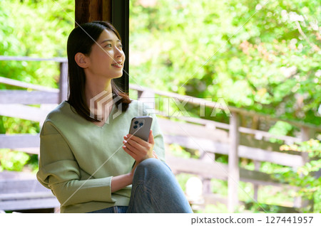 A woman holding a smartphone while gazing at a summer garden on a serene afternoon on a light-filled veranda 127441957