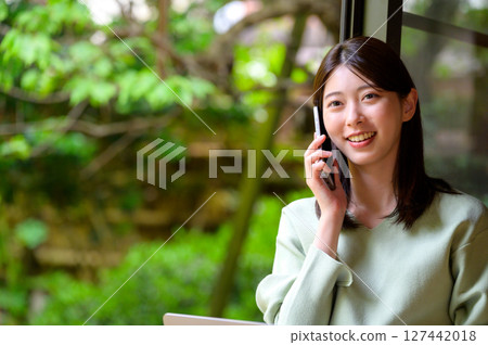 A woman holds her smartphone to her ear indoors and enjoys a phone call with a smile, a lifestyle surrounded by natural summer light A woman holds her smartphone to her ear indoors and enjoys a phone call with a smile, a lifestyle surrounded by natural summer light 127442018