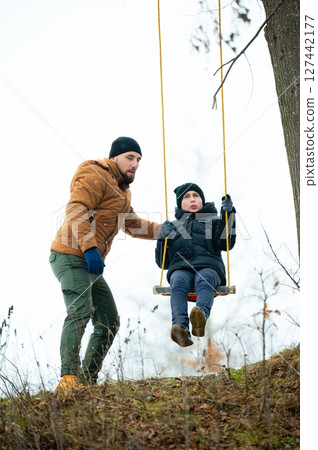 A father helps his son on a swing outdoors in the winter. They are bundled in warm clothing. A father helps his son on a swing outdoors in the winter. They are bundled in warm clothing. 127442177