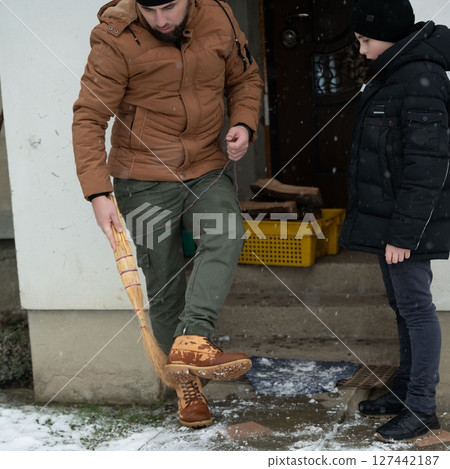 A man playfully sweeps the snow with a broom as a boy watches, creating a lighthearted winter scene outside a house. 127442187