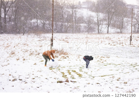 Two individuals construct a snowman during a snowfall in a snowy field. Two individuals construct a snowman during a snowfall in a snowy field. 127442190