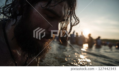 Man with beard emerges from water during spiritual baptism ceremony at sunset with gathered people in the background 127442343