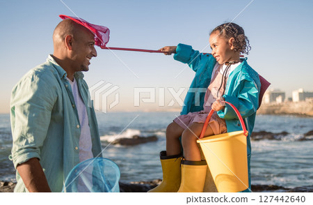 Family fun, girl and father at a beach for learning, playing and collect seashell along the sea, happy and smile against blue sky. Happy family, water and child with dad at the ocean, relax and play 127442640