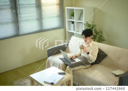 Young man working remotely from home on a sofa with laptop and holding a coffee cup 127442668