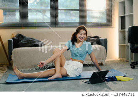 Smiling woman stretches on mat while watching virtual fitness class on digital tablet at home 127442731