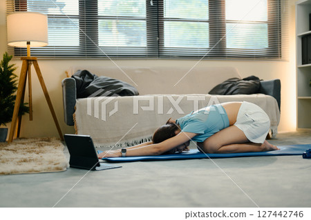 Smiling woman stretches on mat while watching virtual fitness class on digital tablet at home Smiling woman stretches on mat while watching virtual fitness class on digital tablet at home 127442746