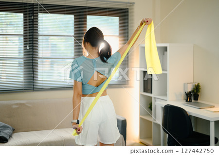 Young woman using a yellow resistance band to stretch her arms during a home workout 127442750