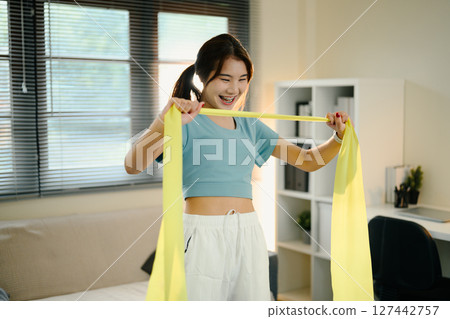 Young woman using a yellow resistance band to stretch her arms during a home workout 127442757