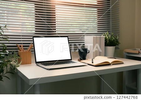Modern home office workspace with a blank laptop screen, open notebook, and coffee mug 127442768